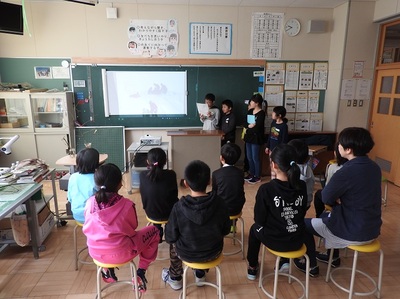 写真が投影された黒板に向かって座る３年生とその前で発表する４年生